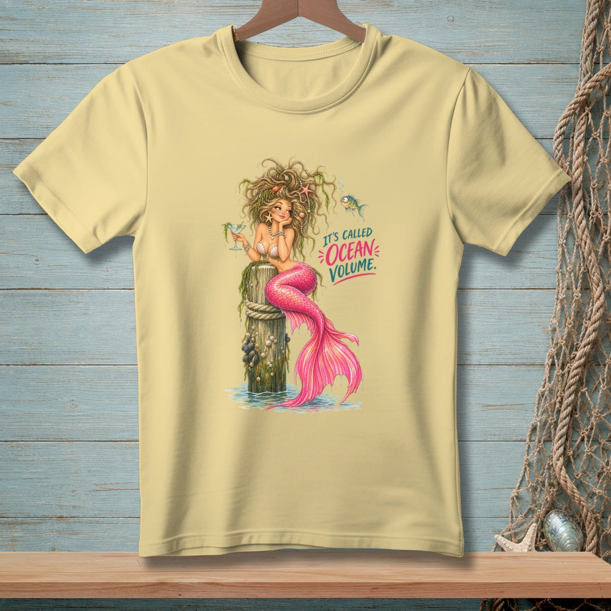 Yellow t-shirt with a cartoon mermaid sitting on a pier, holding a drink. Text reads "IT'S CALLED OCEAN VOLUME."