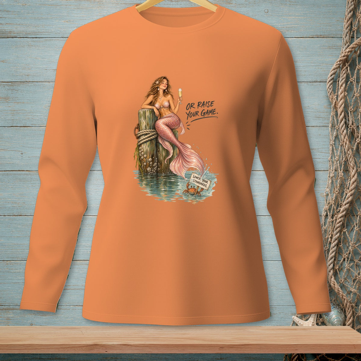 Orange long-sleeve shirt with mermaid and crab graphic, reads "Or Raise Your Game" and "Lower Your Standards".