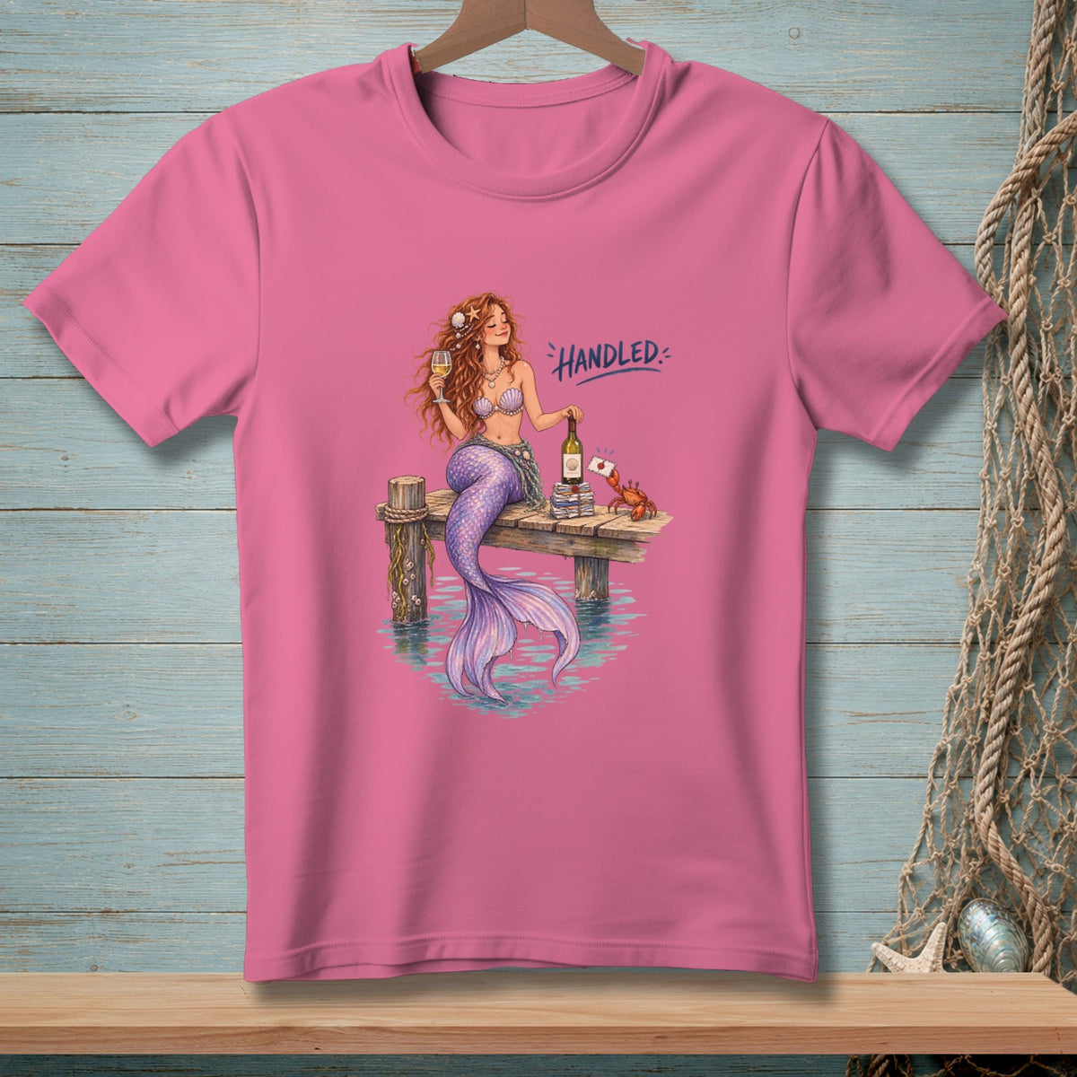 Pink t-shirt with mermaid and crab on a dock, text reads "Handled."