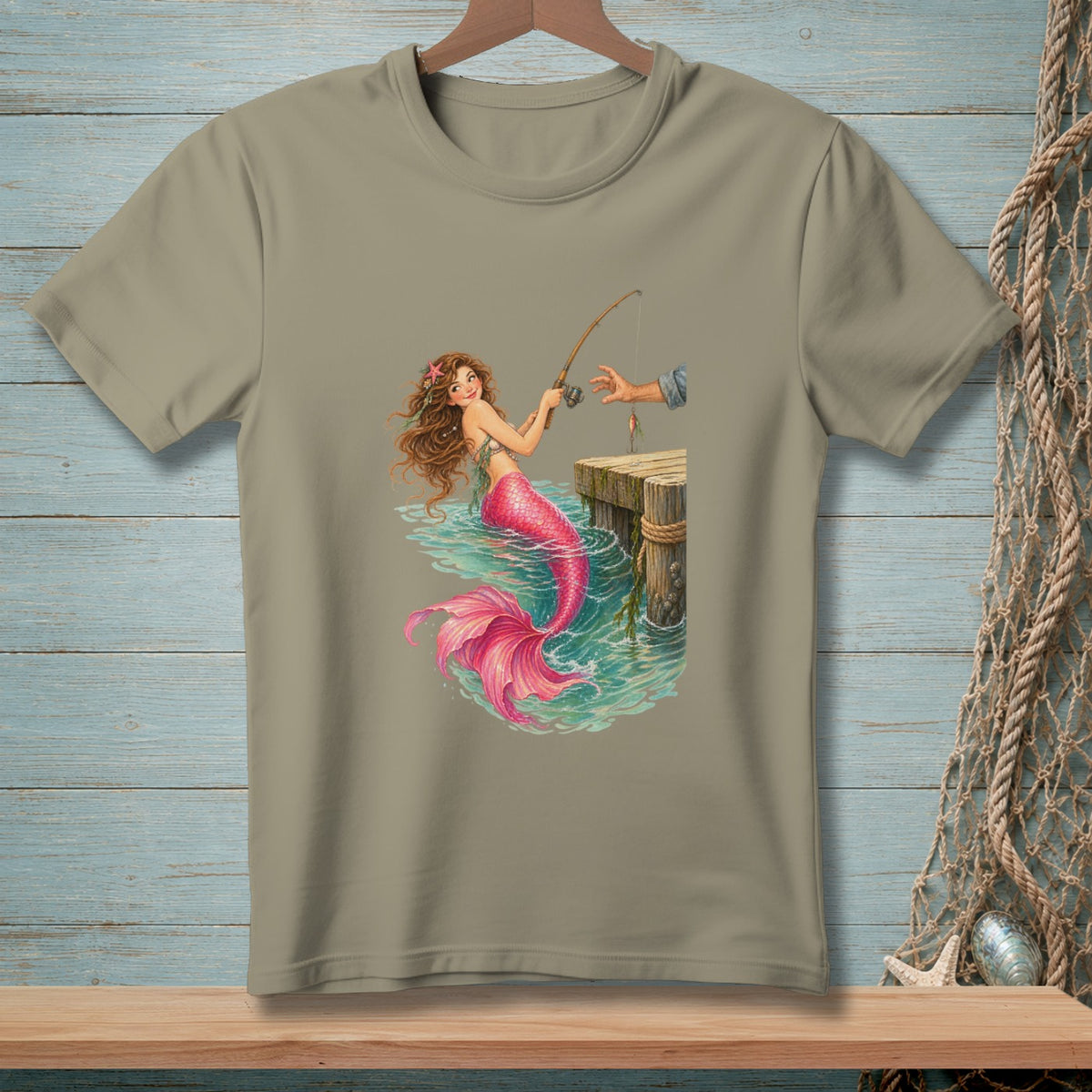 T-shirt graphic of a mermaid fishing from a dock with a hand holding a fishing rod.