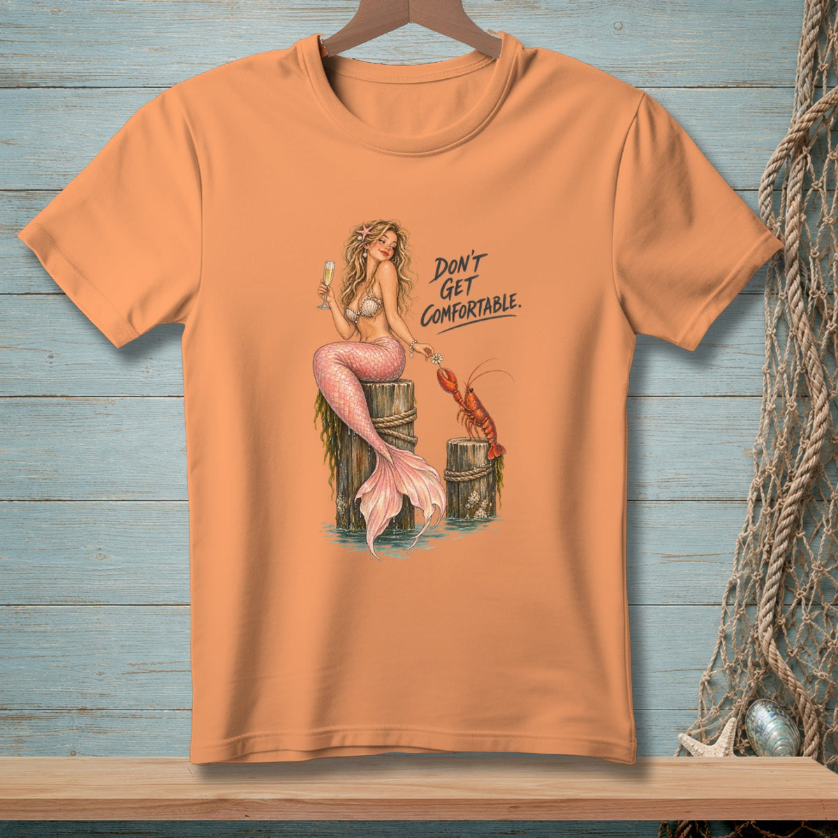 Peach t-shirt with mermaid holding champagne, lobster, and text "DON'T GET COMFORTABLE."