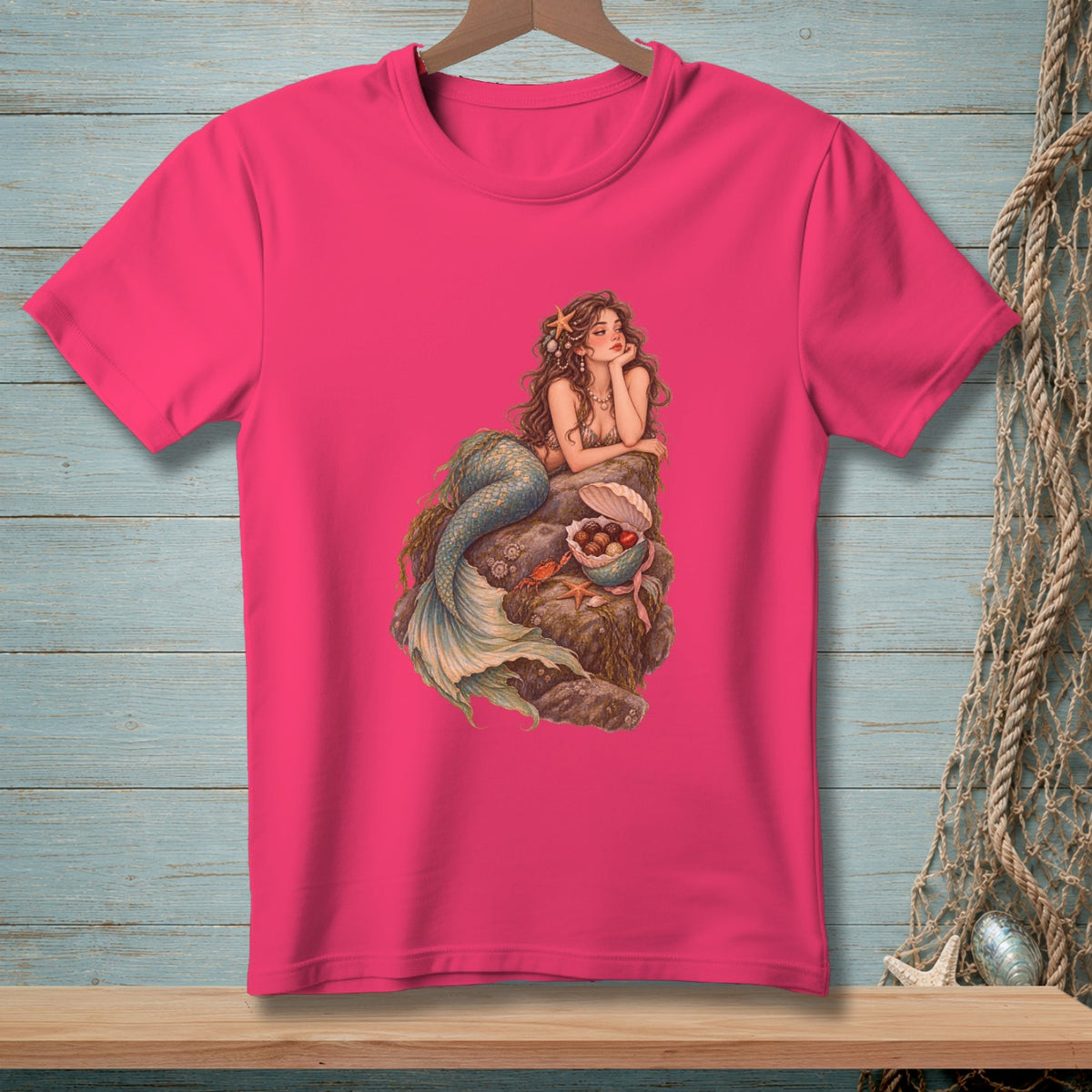 Pink t-shirt with mermaid art featuring a mermaid on rocks surrounded by shells and starfish.