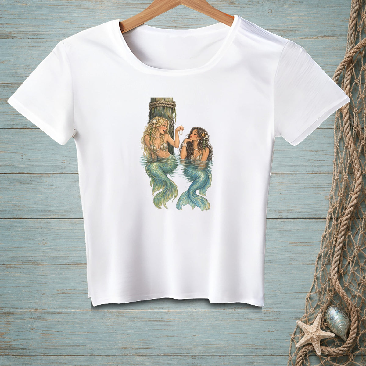 White t-shirt with illustration of two mermaids by a wooden post in water.