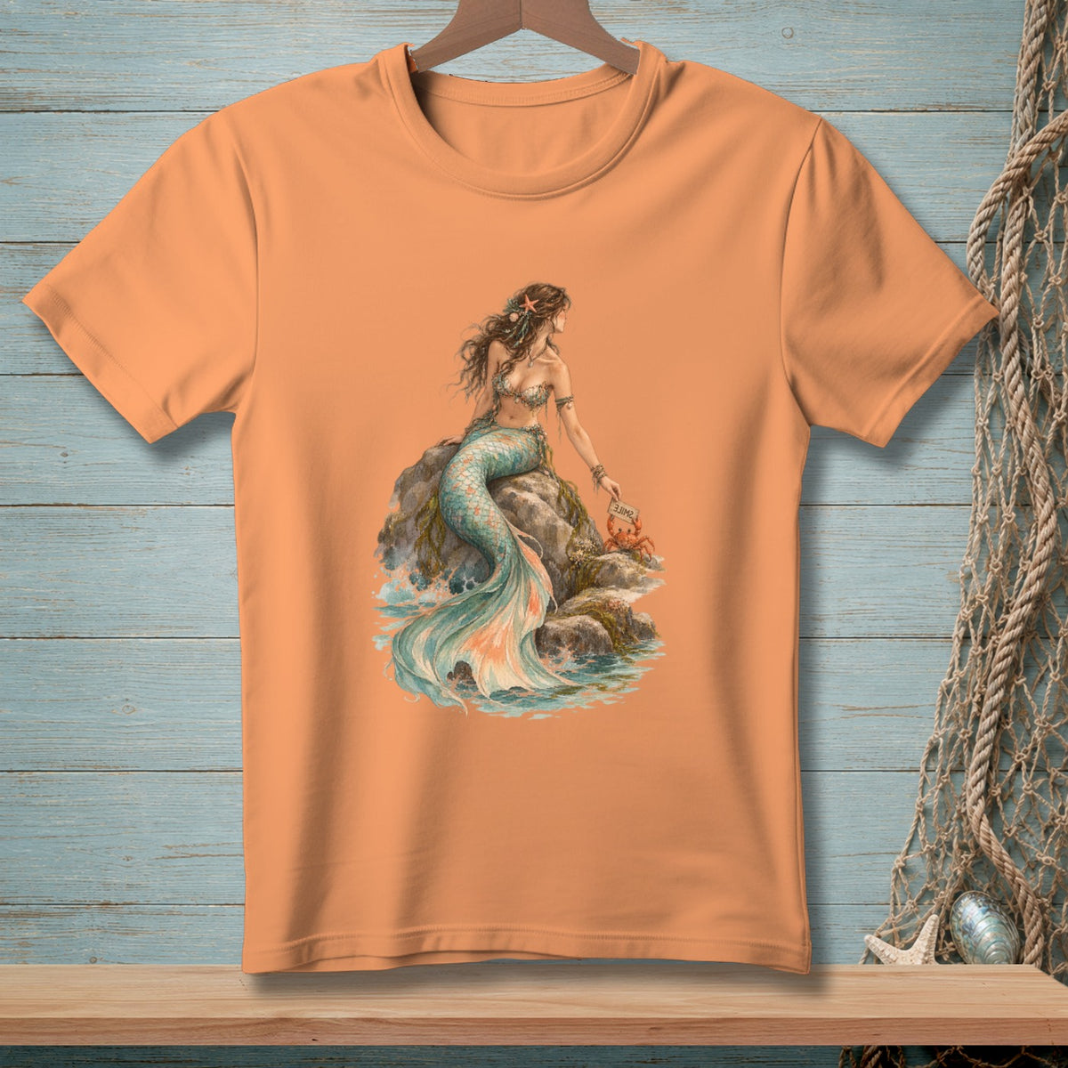Peach t-shirt with illustration of a mermaid on rocks with a crab and a sign.