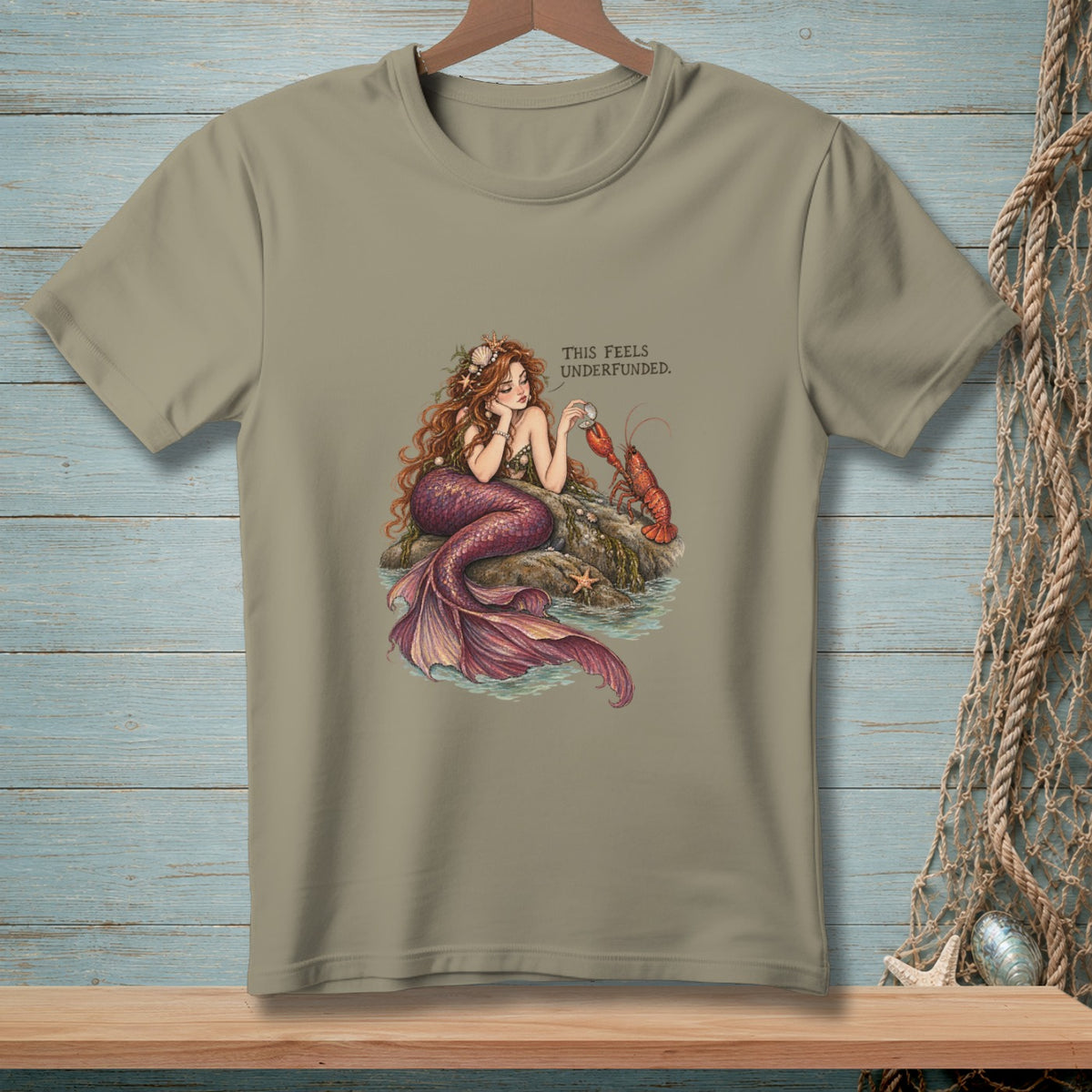 T-shirt graphic: Mermaid resting on rocks with a lobster, text reads "This Feels Underfunded."