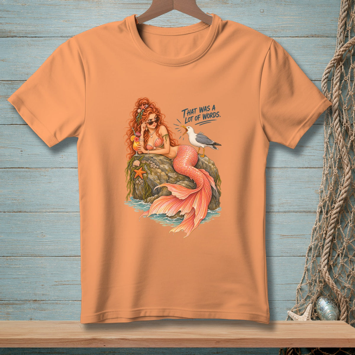Peach t-shirt with graphic of a mermaid on a rock with a seagull and text.