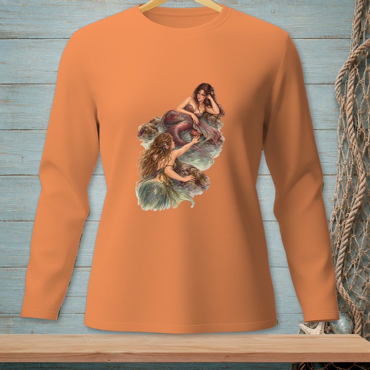 Orange long-sleeve shirt with a graphic of two mermaids on rocks near water.