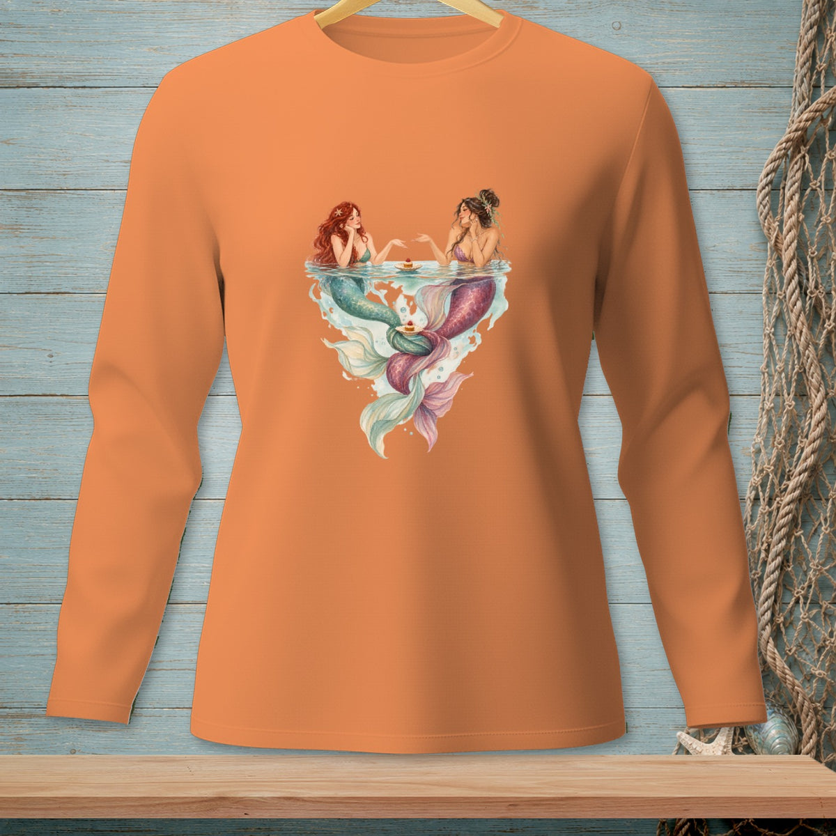 Two mermaids sharing cupcakes in a heart-shaped splash on an orange shirt.