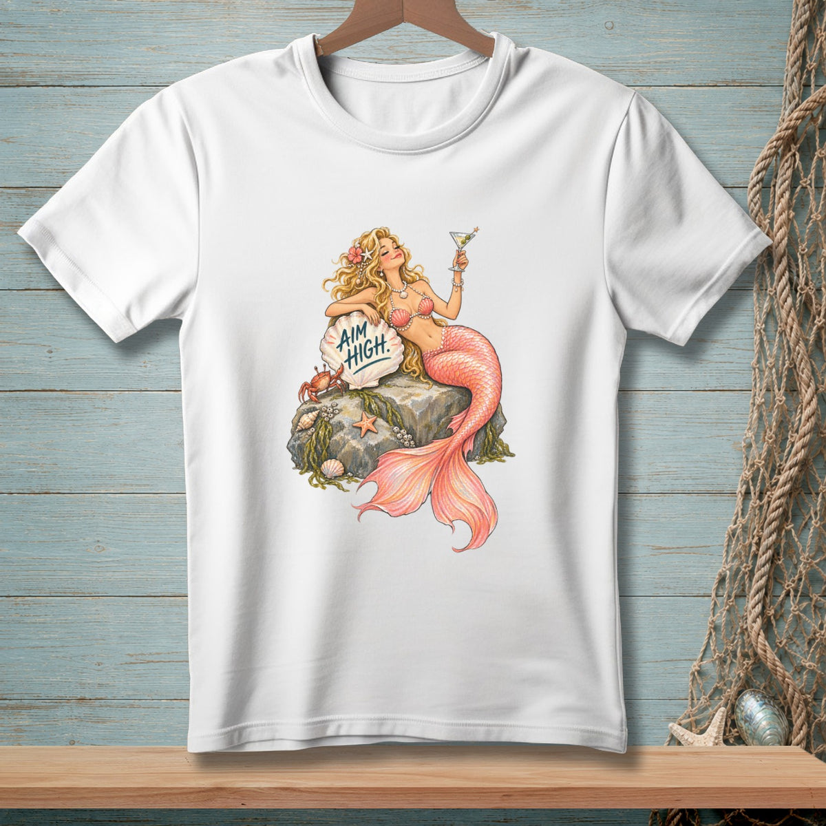 White t-shirt with a drawing of a mermaid holding a martini, resting on a rock with "Aim High" on a shell.