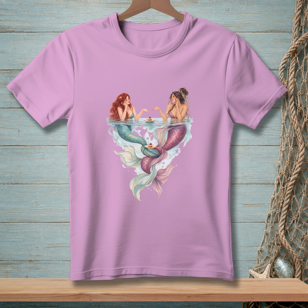 Two mermaids with colorful tails sharing cupcakes by the water.