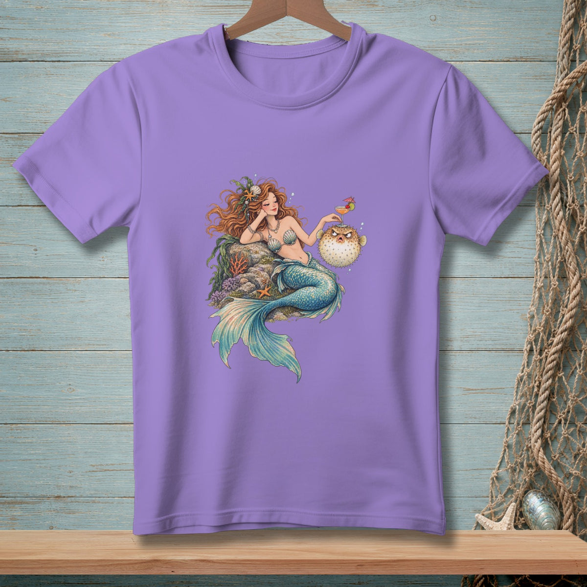 A light purple t-shirt featuring an illustration of a mermaid holding a drink next to a pufferfish.