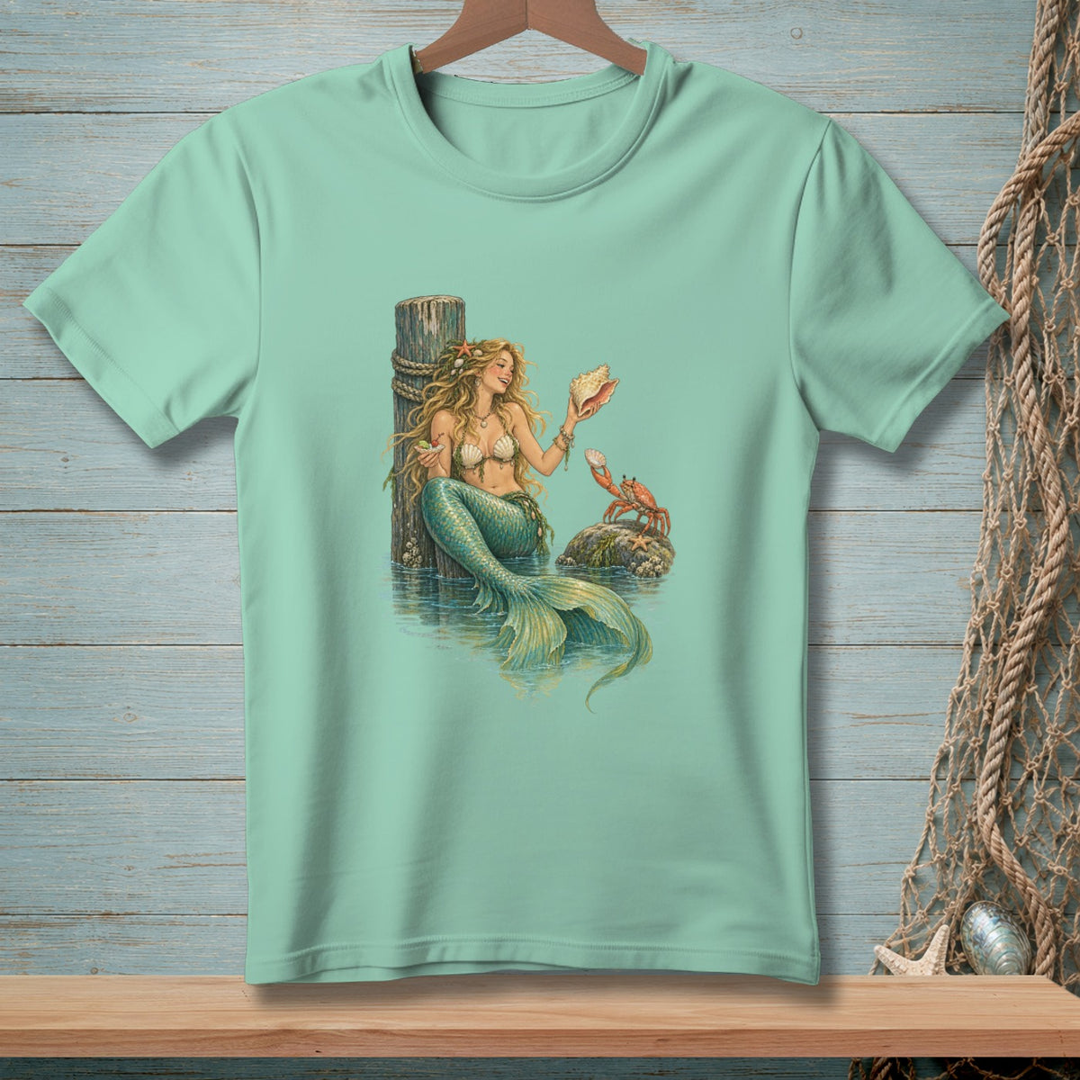 Mint t-shirt with a mermaid, crab, and seashell graphic on a wooden background.