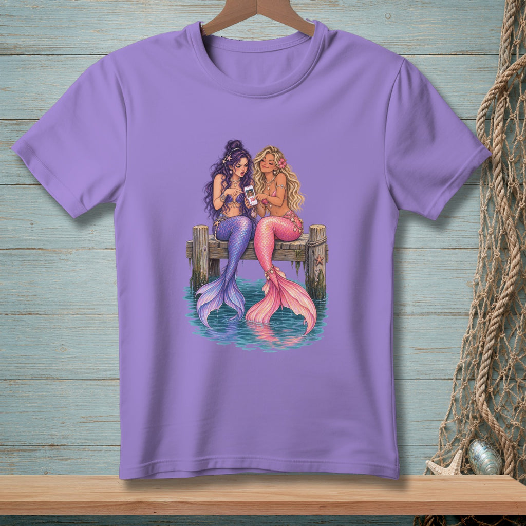 Two mermaids with colorful tails sit on a dock looking at a phone.
