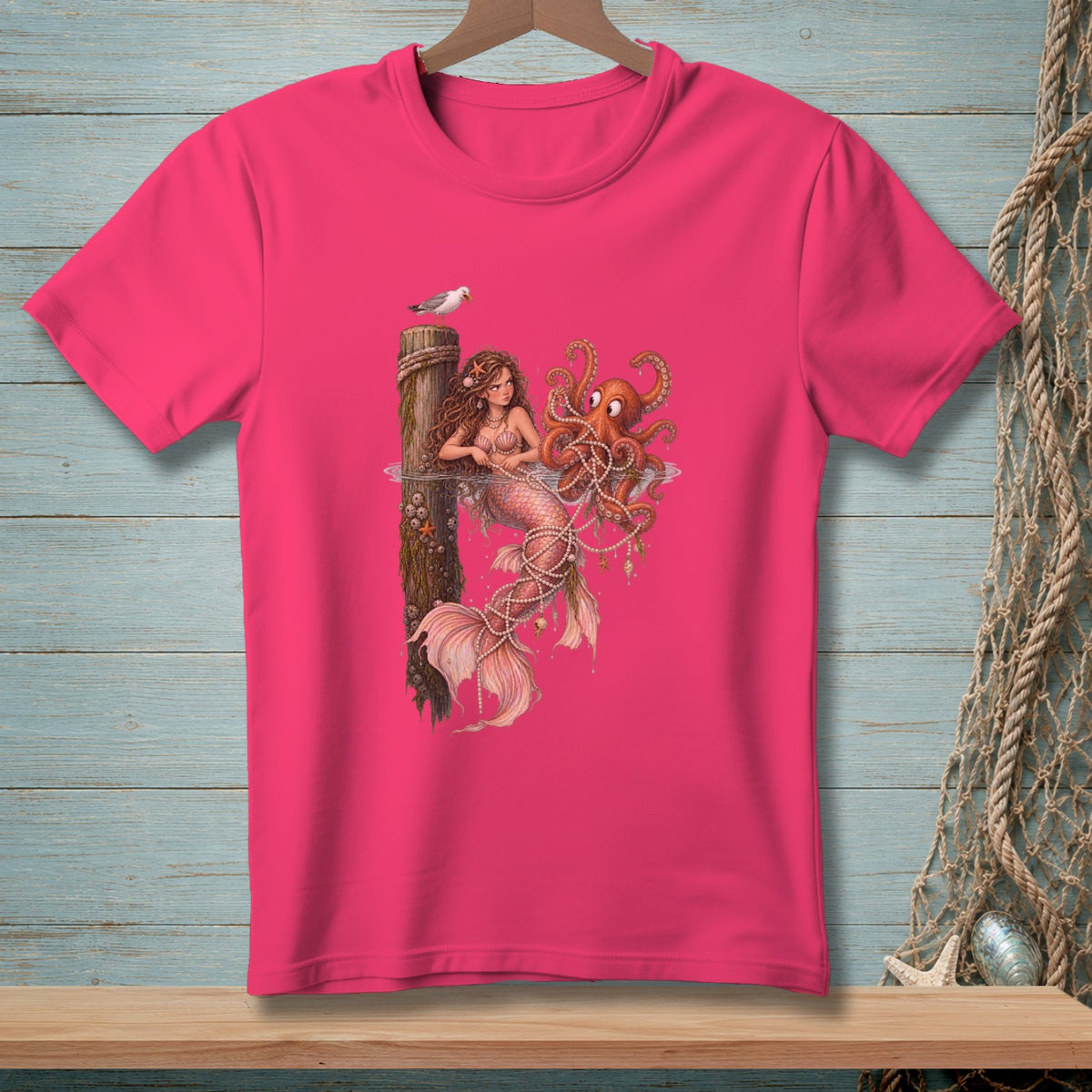 Pink t-shirt with a mermaid, octopus, and seagull graphic on a wooden post.