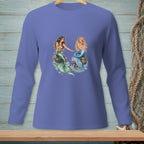 Two mermaids sit on rocks with their tails in the water on a blue long-sleeve shirt.