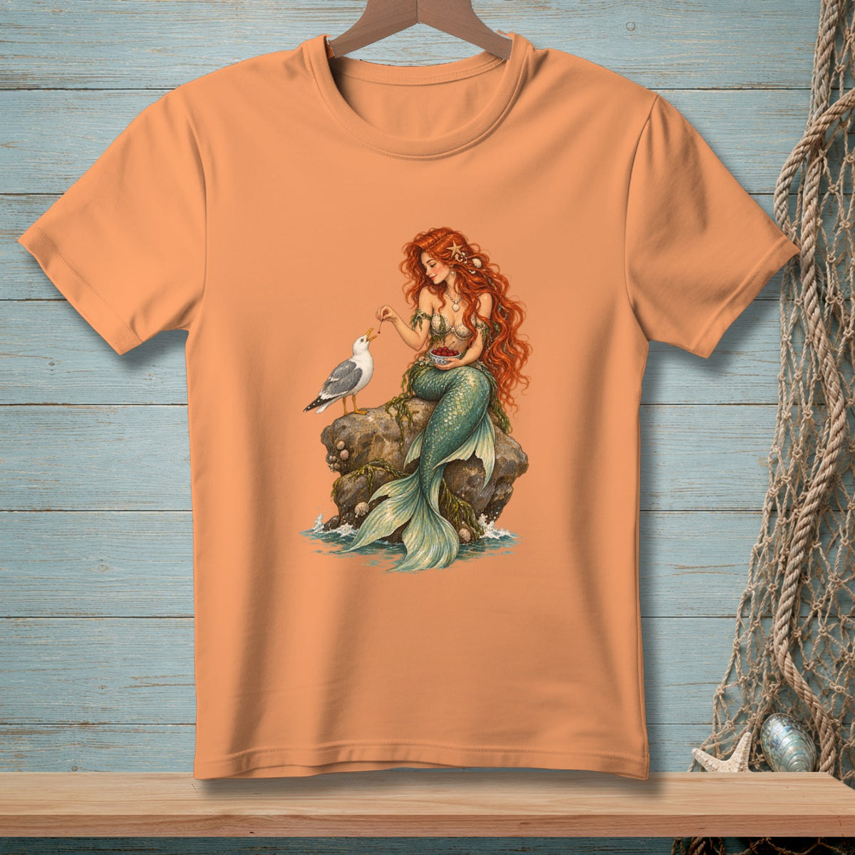 Peach t-shirt with a graphic of a mermaid feeding a seagull on a rock.
