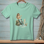 Mint t-shirt with a mermaid sitting on a rock holding a drink, a crab holds a "Be Nice" sign.