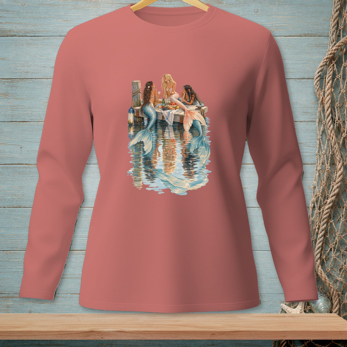 Three mermaids with colorful tails sit on a dock, sharing a meal by the water.