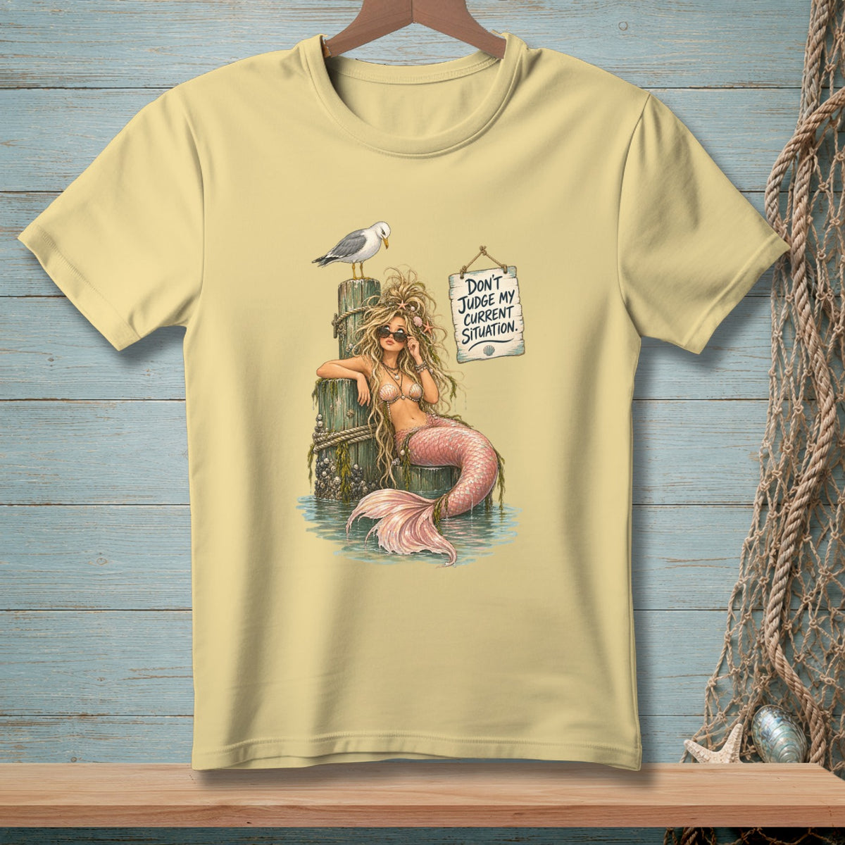 Light yellow t-shirt graphic of a mermaid on a pilothwith a seagull and sign reading "Don't judge my current situation."