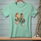Two mermaids sit on rocks by the water with a seagull flying overhead.