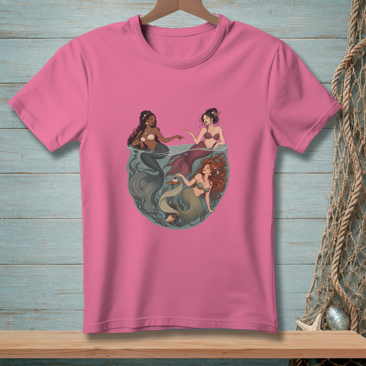 Three mermaids interact within a circular frame on a pink t-shirt.