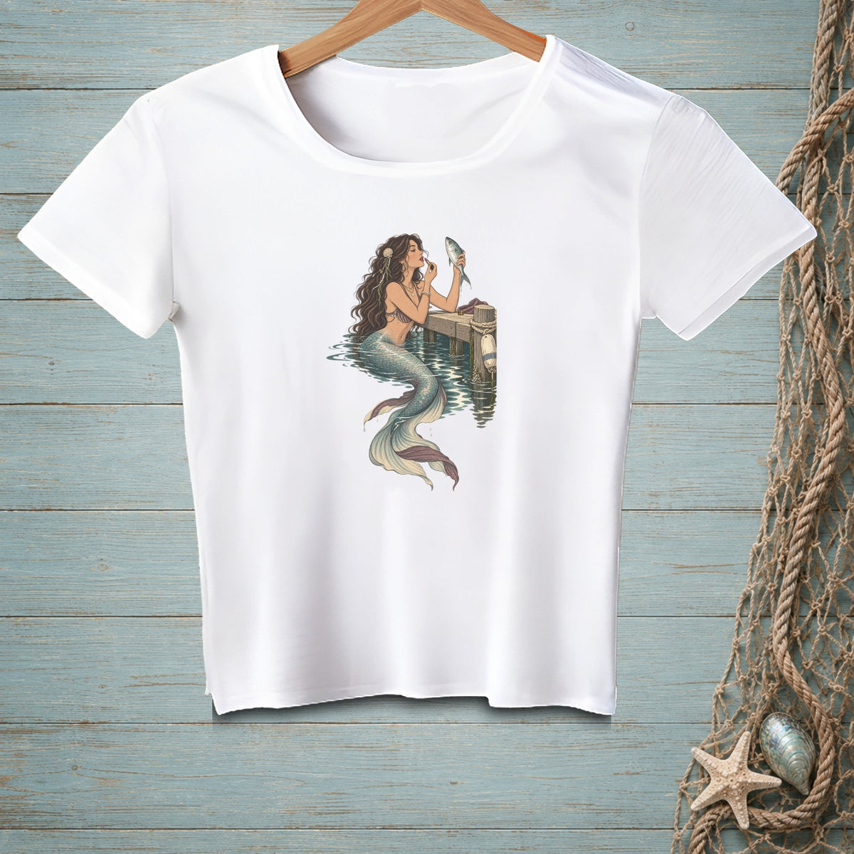 White t-shirt with a mermaid applying makeup to a fish on a pier.