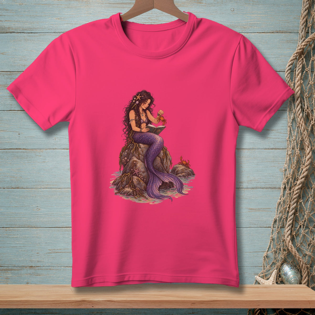 Pink t-shirt with a mermaid reading a book on a rock, holding a crab.