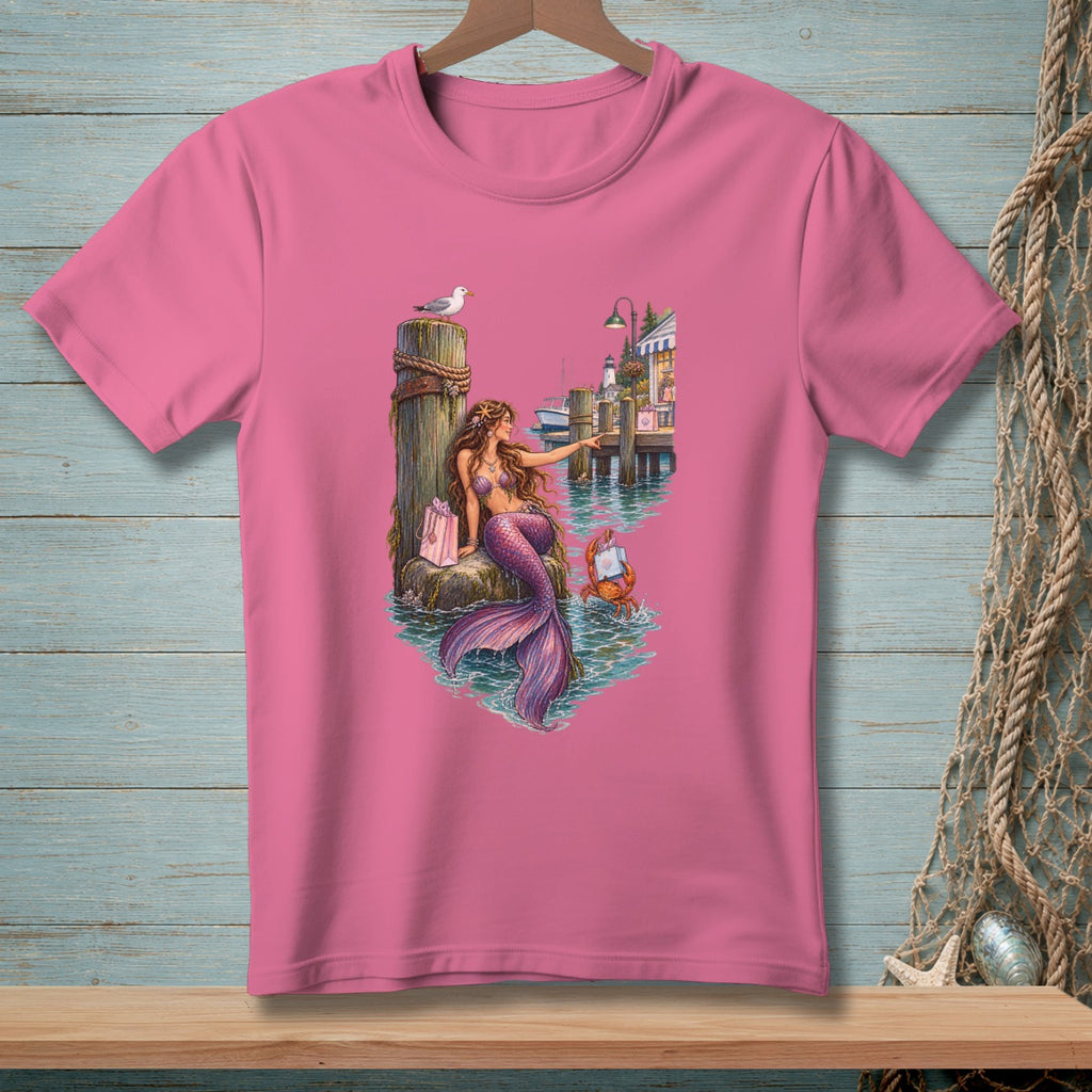 Pink t-shirt with mermaid, crab, gift bag, and waterfront scene illustration.