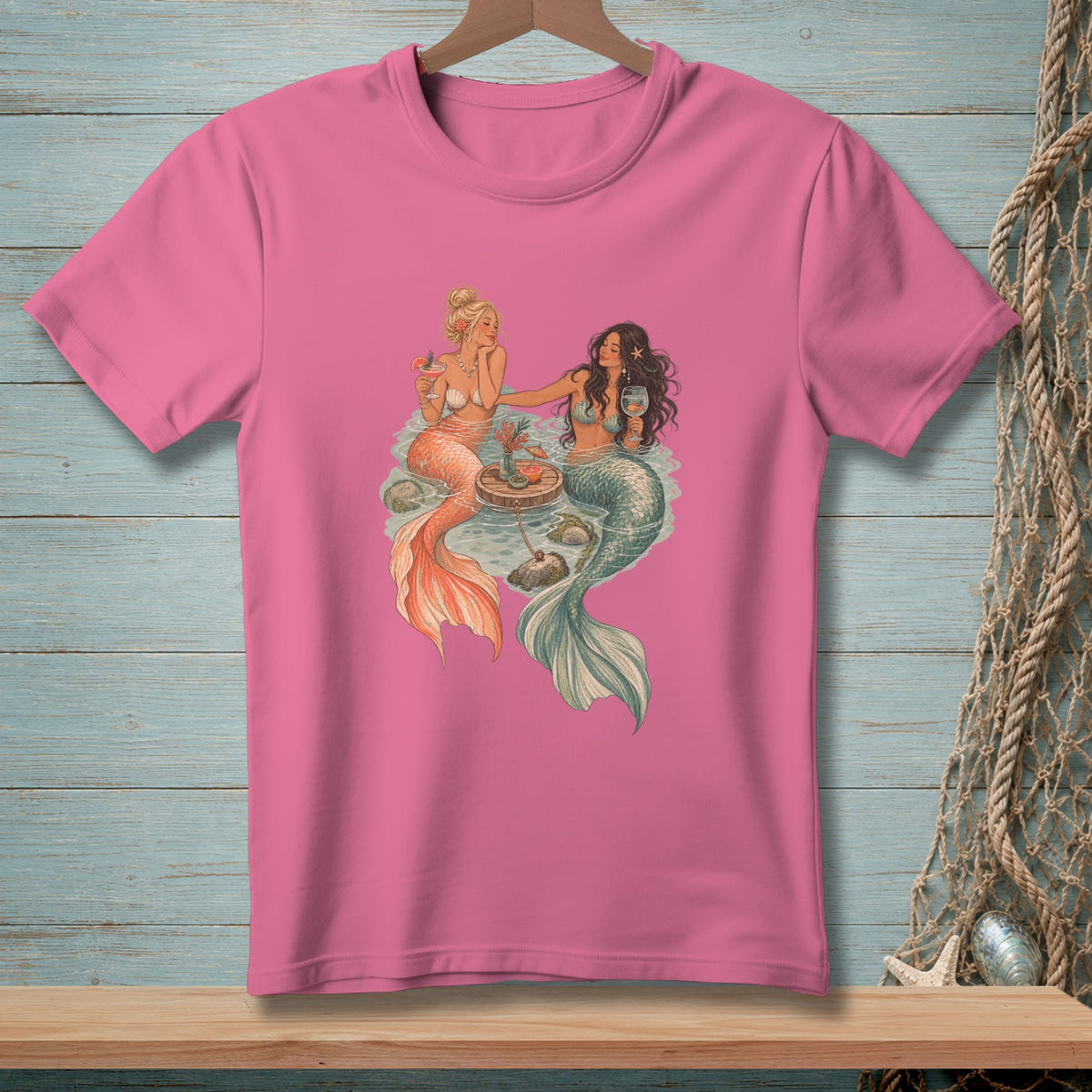 Two mermaids with drinks sit at a small table in the water.