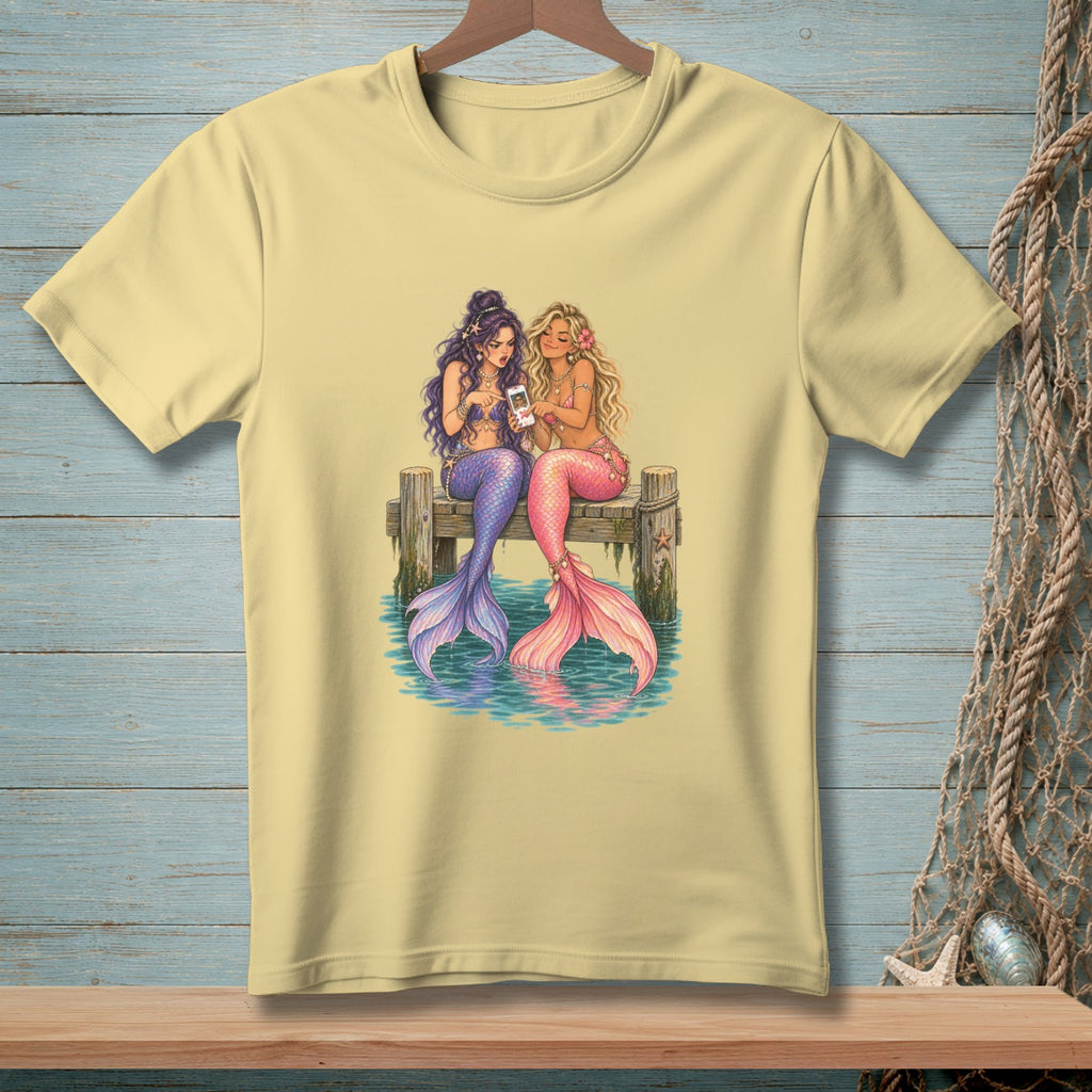 Two mermaids with colorful tails sit on a dock, looking at a smartphone.
