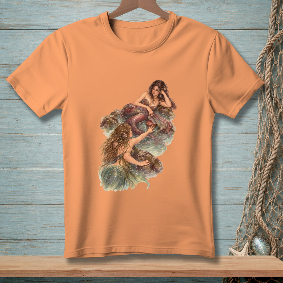 Orange t-shirt featuring an illustration of two mermaids on rocks in water.