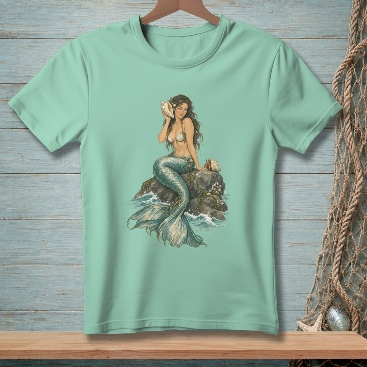 Mint green t-shirt featuring a mermaid holding a conch shell on a rock.