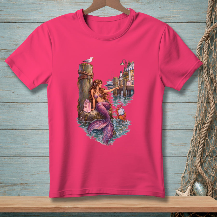 Pink t-shirt featuring a mermaid sitting on a pier with a gift and a seagull.