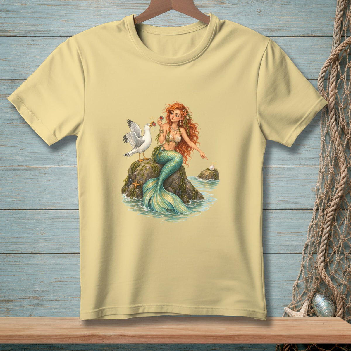 Pale yellow t-shirt with a mermaid and seagull on a rock illustration.