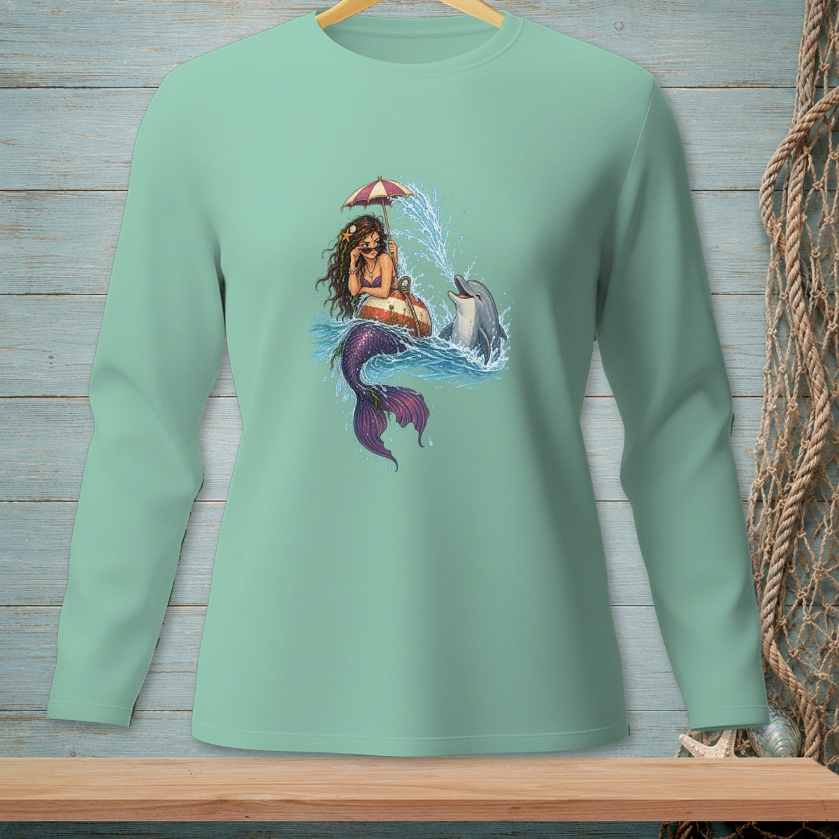 Mermaid under umbrella with dolphin and splashes on a light teal long sleeve shirt.