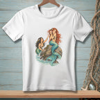 White t-shirt with illustration of two mermaids by rocks and water.