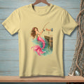Light yellow t-shirt with illustration of a mermaid fishing from a dock.