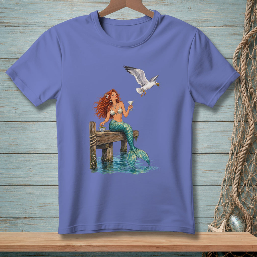 Mermaid with red hair sips a drink on a dock while a seagull flies by.