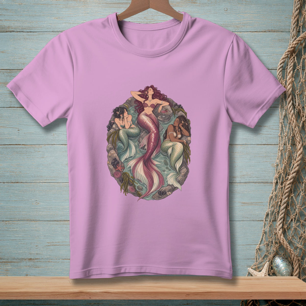 Three mermaids resting in a circular sea-themed graphic on a pink t-shirt.