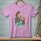 Illustration of a mermaid reading a letter on a pier with a crab and drink.