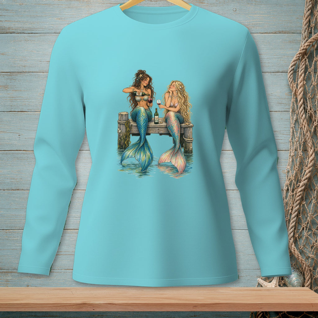 Two mermaids sit on a dock, drinking wine by the water.