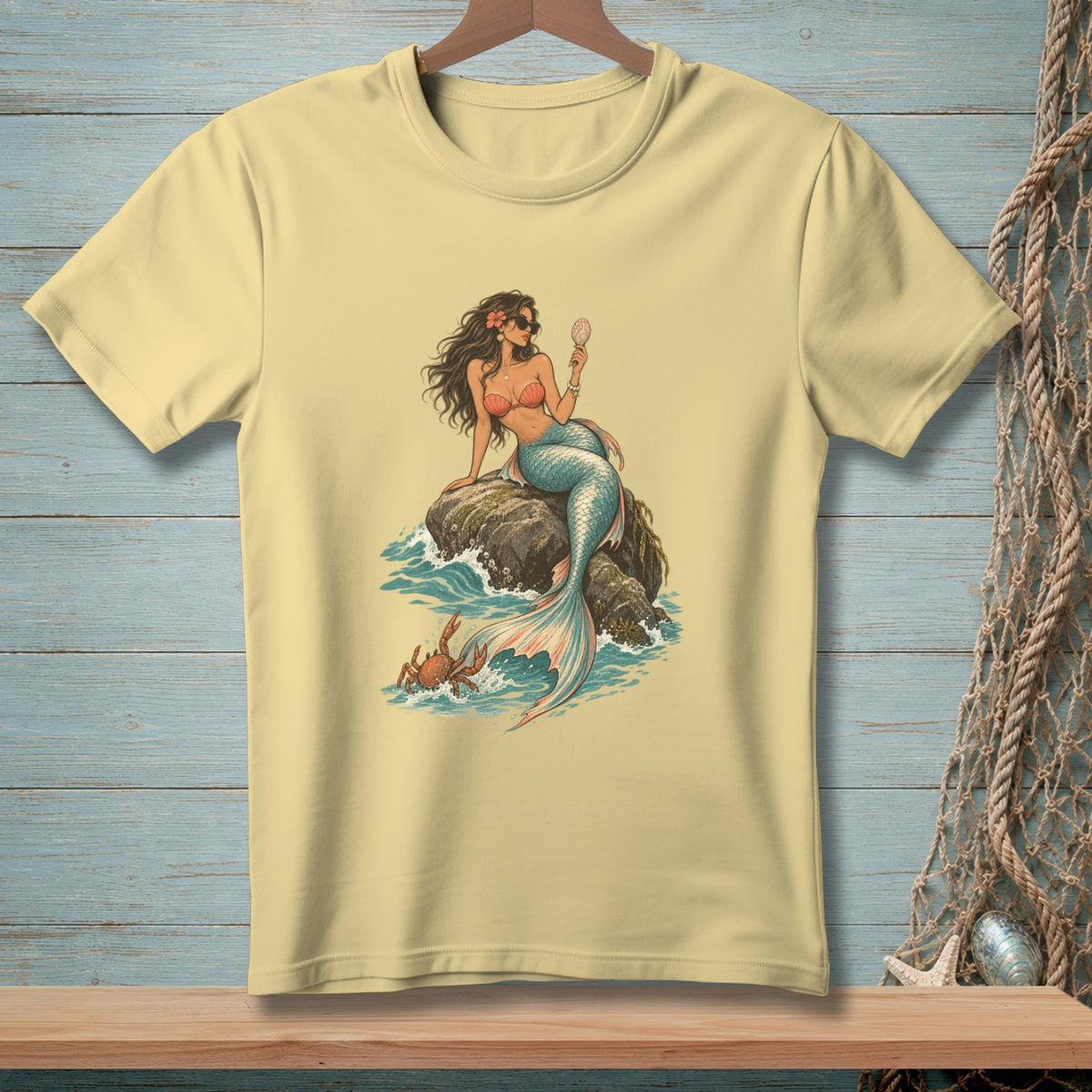 Light yellow t-shirt with a mermaid holding a seashell, sitting on rocks with crabs.