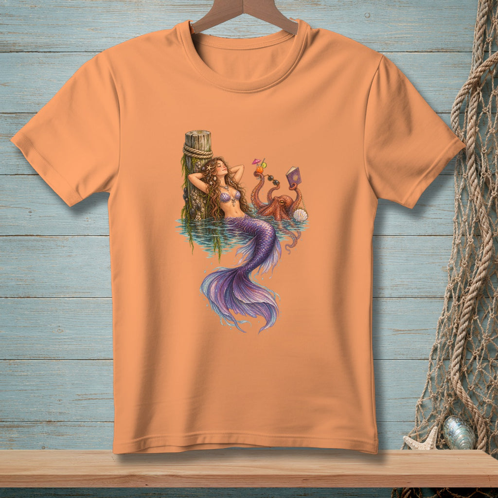 Peach t-shirt with a mermaid and octopus relaxing by a dock.