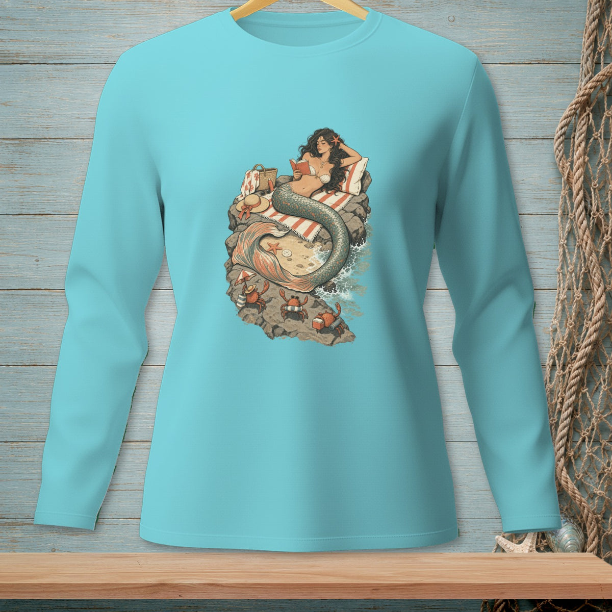 Mermaid reading a book on rocks with crabs and waves, on a blue shirt.