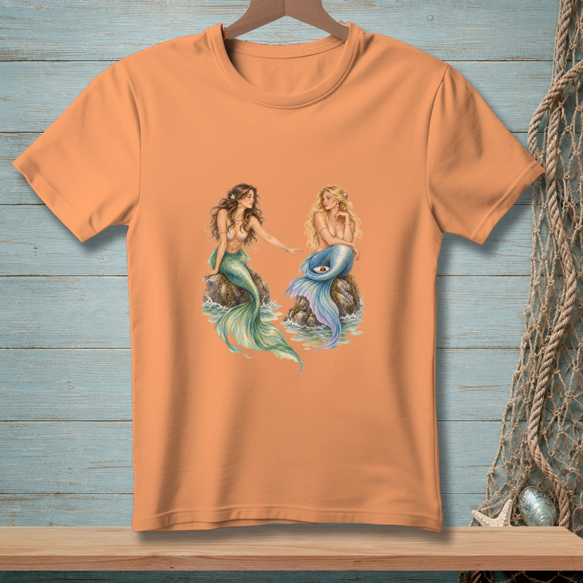 Orange t-shirt with a print of two mermaids sitting on rocks.
