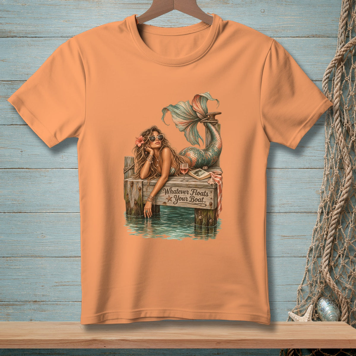 Peach t-shirt with a graphic of a mermaid lounging on a dock, with the text "Whatever Floats Your Boat."