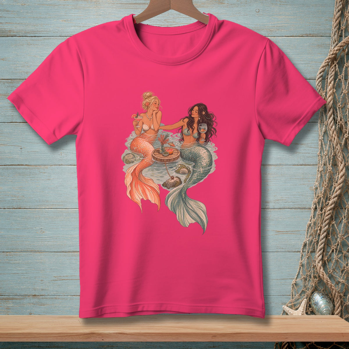 Two mermaids with drinks and a small table with fruit on a pink t-shirt.