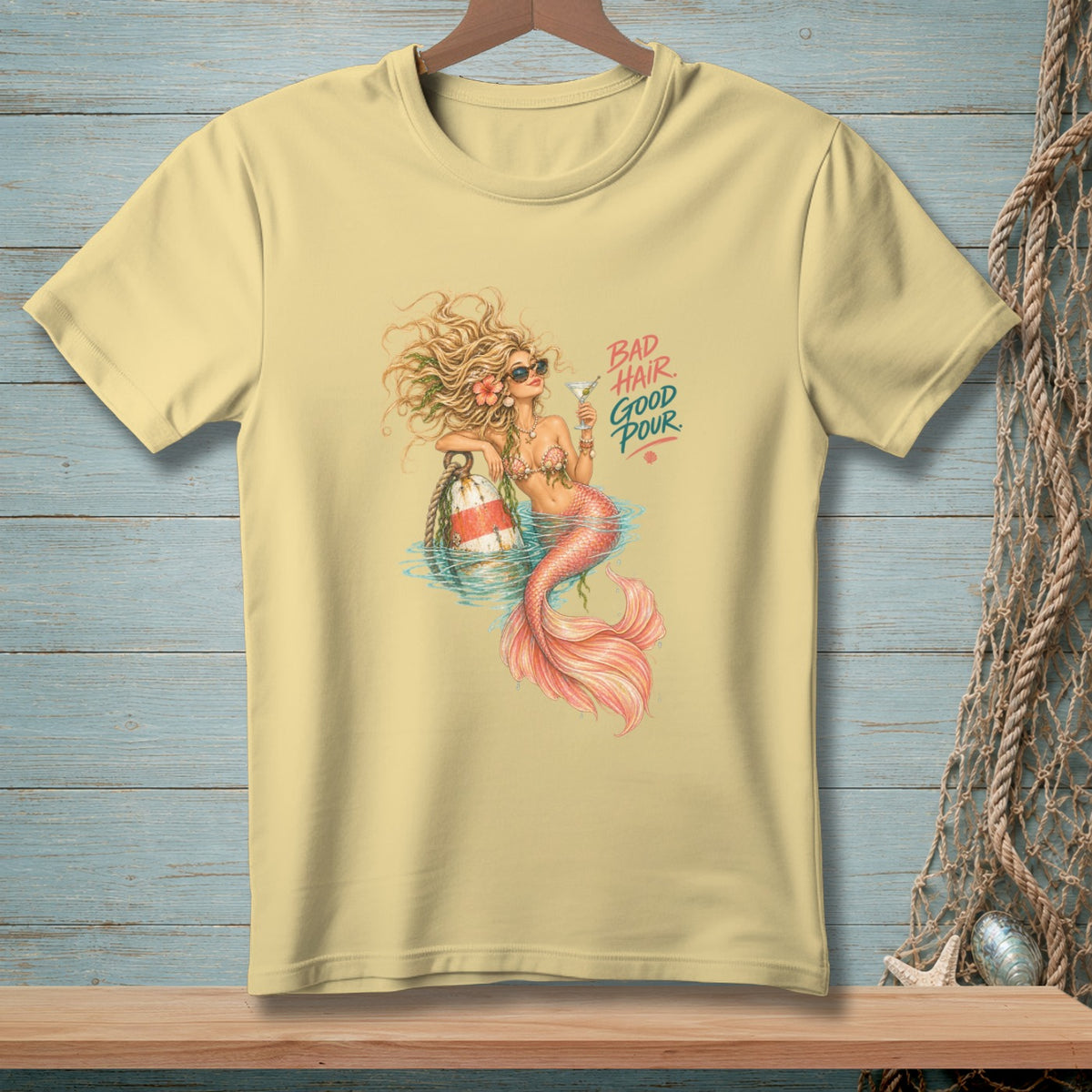 Light yellow t-shirt with a mermaid holding a cocktail and text that reads "BAD HAIR GOOD POUR".
