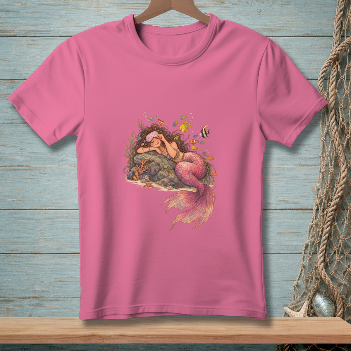 Pink t-shirt with illustration of a sleeping mermaid surrounded by colorful fish and coral.