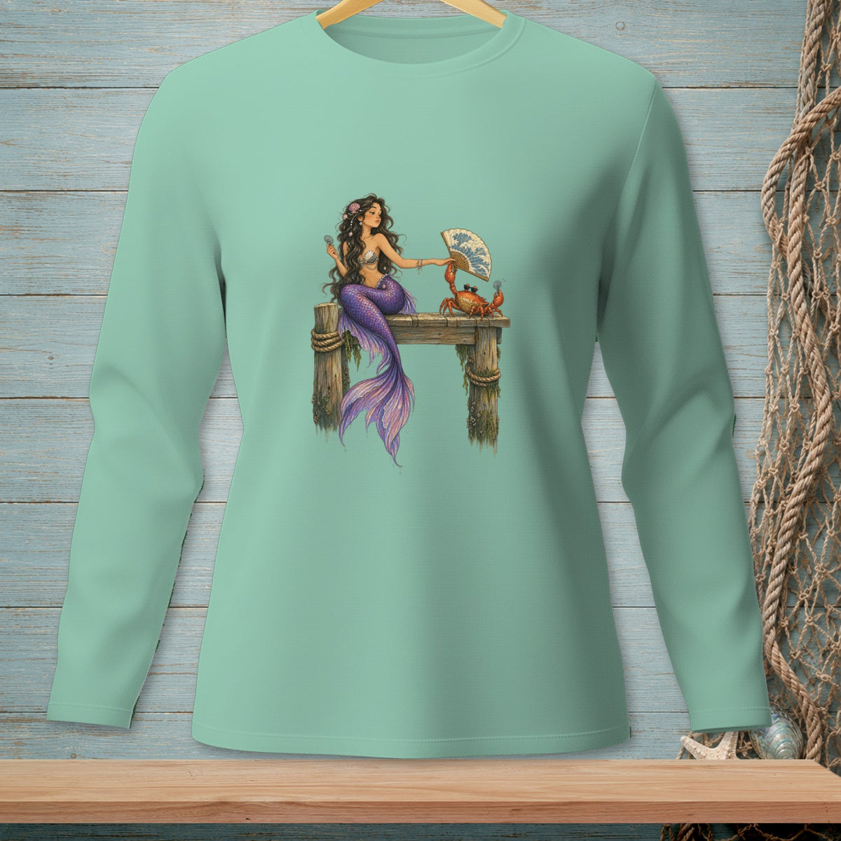 Mermaid with crab holding a fan on a wooden pier graphic on a light green long-sleeved shirt.